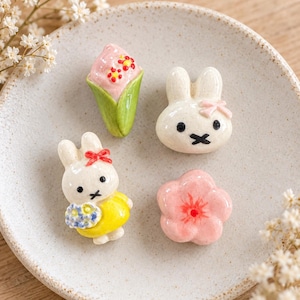 May include: Four small ceramic decorative figures including a pink ice cream cone with red flowers, a white rabbit head with a pink bow, a white rabbit figure wearing a yellow dress and holding blue flowers, and a five-petaled pink flower with a red center.
