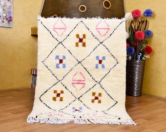 May include: Cream-colored, rectangular rug with a geometric diamond pattern. The design features black outlines, and colorful accents in pink, blue, and brown. The rug has a fringed edge and is displayed indoors.