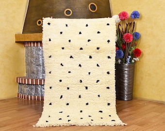 May include: Cream-colored, rectangular rug with scattered black spots. The rug has a shaggy texture and is displayed in front of a fireplace. The rug is a decorative floor covering.