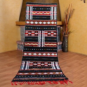 May include: Black and red patterned rug with geometric designs. The rug has a fringe border and is made of wool.