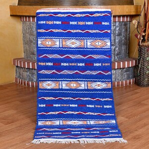 May include: A rectangular blue rug with geometric patterns in white, orange, and red. The rug features a central design of diamond shapes and horizontal bands with zigzag and diamond patterns. The rug has a fringed edge.
