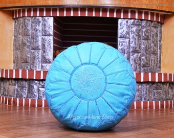 May include: A light blue leather pouf with a decorative stitched pattern and a white embroidered design. The pouf is sitting on a wooden floor in front of a brick fireplace.