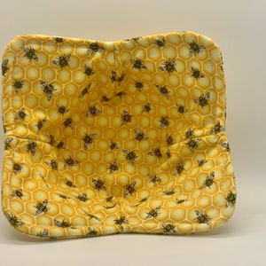 May include: A yellow bowl cozy with a honeycomb pattern and scattered bee illustrations. The cozy is square-shaped with rounded corners and a quilted texture. The interior is a solid yellow color.