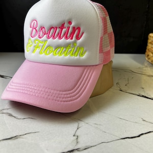 May include: A white foam front trucker hat with a light pink brim and a pink and white checkered mesh back. The front features embroidered text in pink and neon yellow that reads Boatin & Floatin in a script font with a raised texture.