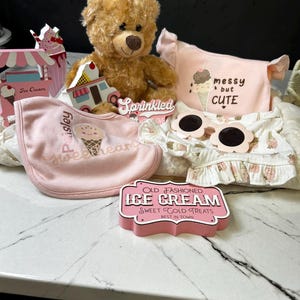 Könnte beinhalten: Ein brauner Teddybär, ein rosa Lätzchen mit Eiscreme-Grafik und dem Text Paisley sweetheart, ein rosa Oberteil mit dem Text messy but CUTE, blumenförmige Sonnenbrillen und Holzschilder mit der Aufschrift Sprinkled und OLD FASHIONED ICE CREAM.