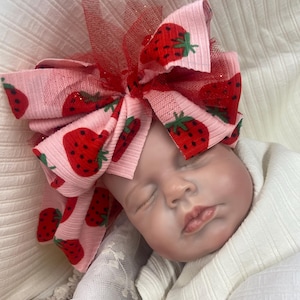 Peut inclure: Un grand nœud pour cheveux en tissu côtelé rose clair avec un motif répétitif de fraises rouges. Le nœud présente des couches de tulle rouge pailleté insérées au centre et est porté sur la tête d'une poupée nourrisson endormie.