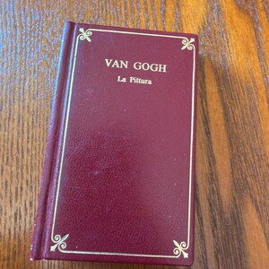 May include: A small, burgundy-colored book with gold trim and the title "VAN GOGH La Pittura" in gold lettering. The book has decorative corner accents and is set against a wooden surface.