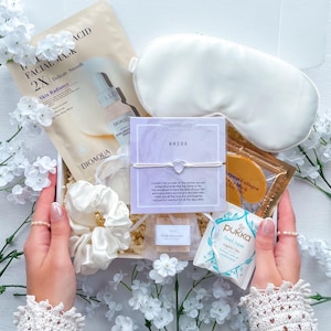 May include: A white gift box containing bridal self-care products. It features a white eye mask, face masks, a bracelet with a heart charm, soap, and a tea bag. The box is decorated with white flowers.