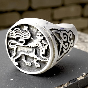 Puede incluir: Un anillo plateado con un diseño detallado de escudo de león. El anillo presenta una cara circular con un emblema de león y corona, y un patrón adornado en el lateral. El anillo está sobre una superficie oscura.