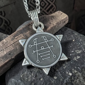 May include: A silver-toned pendant necklace featuring a circular emblem with intricate symbols and a six-pointed star frame. The pendant hangs from a silver chain. The background includes a wooden stick and a dark, textured surface.