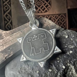 May include: A silver-colored pendant necklace featuring a circular emblem with a geometric design and a starburst border. The pendant hangs from a silver chain. The background includes wood and stone.