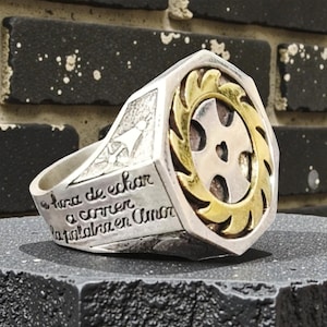 Könnte beinhalten: Silberfarbener Ring mit goldenem Akzent, der ein Kreuzdesign aufweist. Der Ring hat gravierten Text auf dem Band. Der Ring wird auf einer dunklen Oberfläche mit Backsteinhintergrund präsentiert.