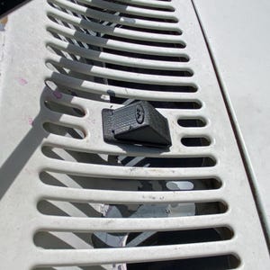 May include: A black plastic latch with a small round button on the top. The latch is attached to a white metal surface with a series of parallel lines.
