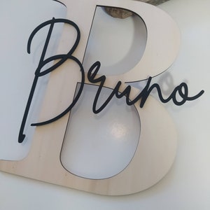 May include: A large, light-colored wooden letter B with a smooth finish, featuring the name Bruno in a black, cursive, script-style font layered directly over the center of the letter. The design is flat and cut from thin, solid material.