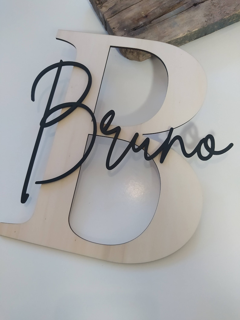 May include: A large, light-colored wooden letter B with a smooth finish, featuring the name Bruno in a black, cursive, script-style font layered directly over the center of the letter. The design is flat and cut from thin, solid material.