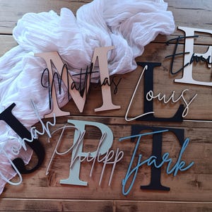 May include: A collection of custom wooden wall letters featuring large serif initials layered with smaller cursive names. The set includes the names Jonah, Matthias, Philipp, Louis, Tjark, and Emily in various colors including black, white, and blue.