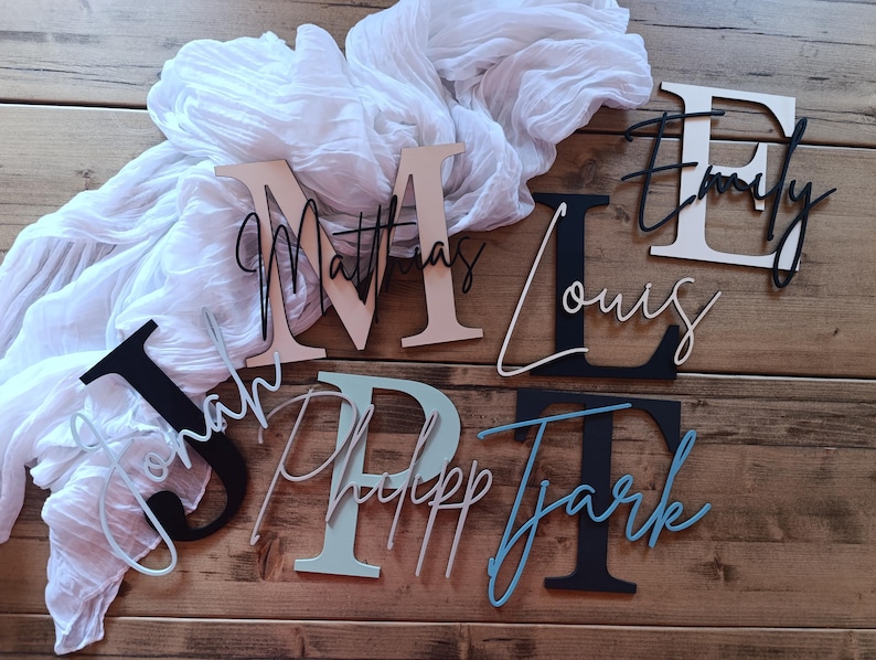 May include: A collection of custom wooden wall letters featuring large serif initials layered with smaller cursive names. The set includes the names Jonah, Matthias, Philipp, Louis, Tjark, and Emily in various colors including black, white, and blue.
