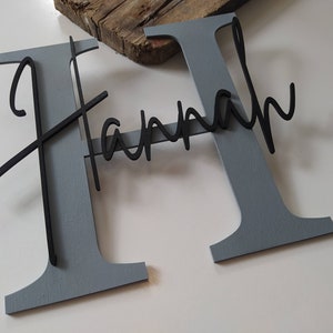 May include: A large, gray, block-letter H with the name Hannah written in a black, cursive, script-style font layered across the front. The name is attached to the center of the letter, creating a dimensional effect with contrasting colors.