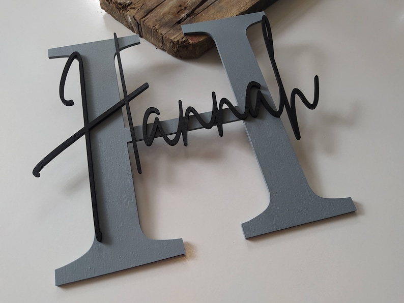 May include: A large, gray, block-letter H with the name Hannah written in a black, cursive, script-style font layered across the front. The name is attached to the center of the letter, creating a dimensional effect with contrasting colors.
