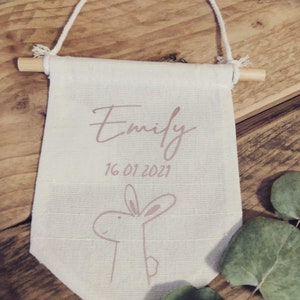 Könnte beinhalten: Ein weißer Canvas-Banner mit einer rosa Hasenillustration und dem Text "Emily 16 01 2021" hängt an einem Holzstab.