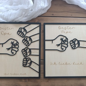 Könnte beinhalten: Zwei gerahmte Holztafeln mit schwarzen Umrissen und Text. Eine sagt "Bester Opa" und "Wir lieben dich" mit einem Faust-Design und Namen. Die andere sagt "Bester Papa" und "Ich liebe dich" mit einem Faust-Design und einem Namen.