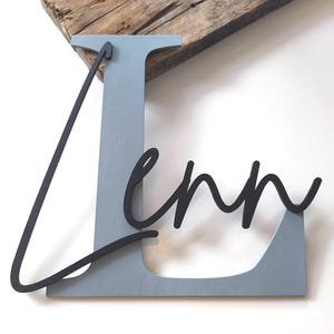 May include: A large, light blue wooden letter L with a smooth finish, featuring the name Lenn in a black, cursive, three-dimensional script font layered over the front of the letter. The piece is constructed from wood and has a matte appearance.