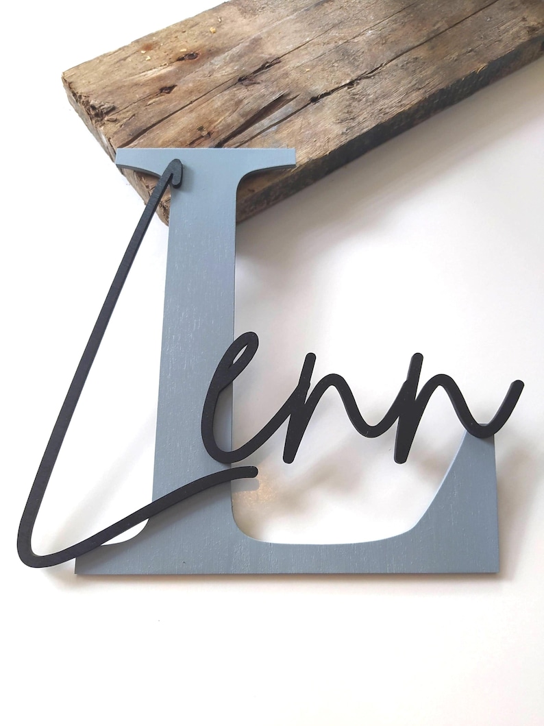 May include: A large, light blue wooden letter L with a smooth finish, featuring the name Lenn in a black, cursive, three-dimensional script font layered over the front of the letter. The piece is constructed from wood and has a matte appearance.