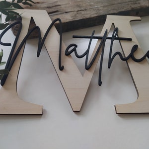 May include: A large, light-colored wooden letter M with the name Matthew written in black cursive script layered over the front. The name is connected to the letter, creating a two-tone decorative sign with a natural wood finish and black text.