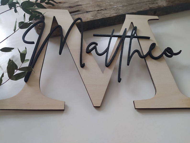 May include: A large, light-colored wooden letter M with the name Matthew written in black cursive script layered over the front. The name is connected to the letter, creating a two-tone decorative sign with a natural wood finish and black text.