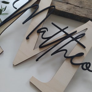 May include: A large, light-colored wooden letter M with a smooth finish, featuring the name "Matheo" in a black, cursive, raised script font layered on top. The black lettering is positioned diagonally across the center of the wooden letter.