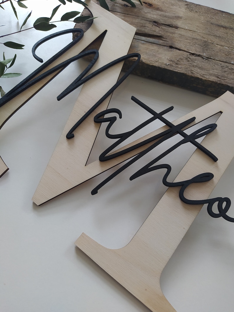 May include: A large, light-colored wooden letter M with a smooth finish, featuring the name "Matheo" in a black, cursive, raised script font layered on top. The black lettering is positioned diagonally across the center of the wooden letter.