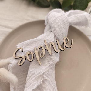 Könnte beinhalten: Ein Namensschild aus Holz mit dem Namen "Sophie" in Schreibschrift liegt auf einer weißen Stoffserviette. Die Serviette ist über einem beigen Teller drapiert, mit Grün im Hintergrund. Ideal für Hochzeiten oder besondere Anlässe.
