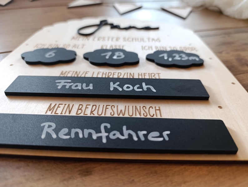 May include: A wooden sign with black chalkboard-style accents. The sign includes the text "June", "Mein Erster Schultag", and "Frau Koch". It also features cloud-shaped cutouts with numbers and measurements, and the word "Rennfahrer".