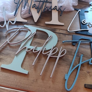 May include: A collection of wooden letters and names featuring various colors including black, white, sage green, and blue. Each piece consists of a large block letter with a cursive name layered on top, such as Matthias, Sarah, Philipp, Louis, and Jaro.