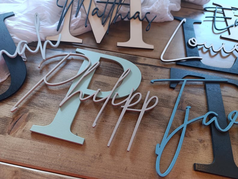 May include: A collection of wooden letters and names featuring various colors including black, white, sage green, and blue. Each piece consists of a large block letter with a cursive name layered on top, such as Matthias, Sarah, Philipp, Louis, and Jaro.