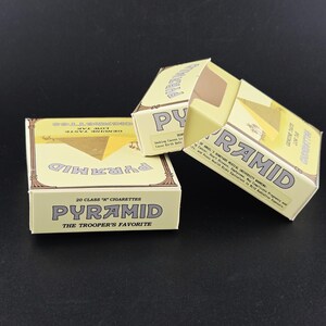 May include: Three light yellow cigarette boxes with the word "PYRAMID" in bold letters. The boxes have a pyramid graphic and text that reads "20 CLASS 'A' CIGARETTES" and "THE TROOPER'S FAVORITE". The boxes are on a dark surface.