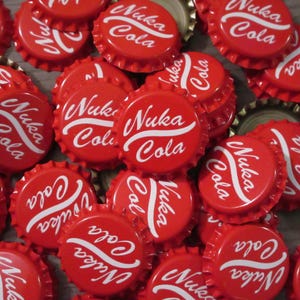 May include: A collection of red metal bottle caps, each featuring the white text Nuka Cola in a stylized script font. The caps are arranged in a dense, overlapping pile, showcasing the circular design and the serrated edges typical of standard bottle closures.
