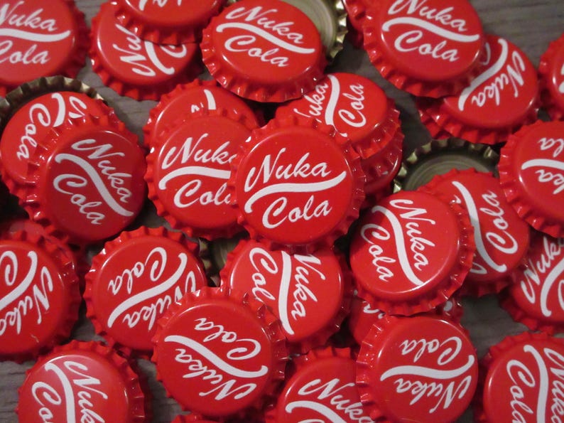May include: A collection of red metal bottle caps, each featuring the white text Nuka Cola in a stylized script font. The caps are arranged in a dense, overlapping pile, showcasing the circular design and the serrated edges typical of standard bottle closures.