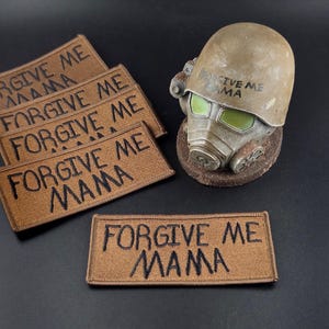 Peut inclure: Quatre écussons rectangulaires brodés marron avec le texte noir FORGIVE ME MAMA, accompagnés d'un casque tactique beige avec un masque à gaz et des lentilles vertes, portant également l'inscription FORGIVE ME MAMA en lettres noires.