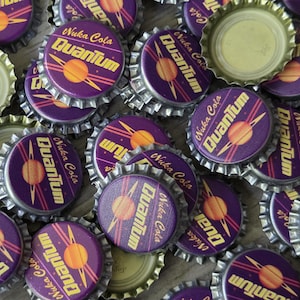 May include: A collection of metal bottle caps featuring a purple background with a yellow planet graphic and the text Nuka Cola Quantum. The caps have a serrated edge and some are flipped over to show the plain metallic interior lining.