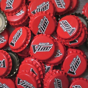 May include: A collection of red metal bottle caps featuring the white and black text "You've got... Vim!" printed on the top. The caps are arranged in a pile, showing both the exterior branded sides and the metallic underside of some caps.