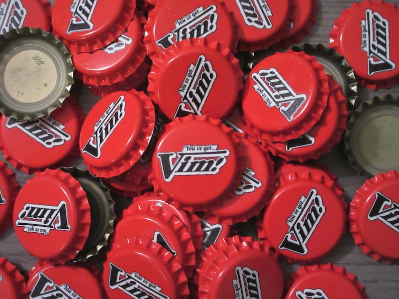 May include: A collection of red metal bottle caps featuring the white and black text "You've got... Vim!" printed on the top. The caps are arranged in a pile, showing both the exterior branded sides and the metallic underside of some caps.