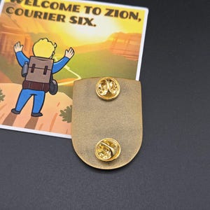 May include: A gold-colored metal pin with two pin backs. The pin is shield-shaped. A card with the text "WELCOME TO ZION, COURIER SIX" and a cartoon character is next to the pin.