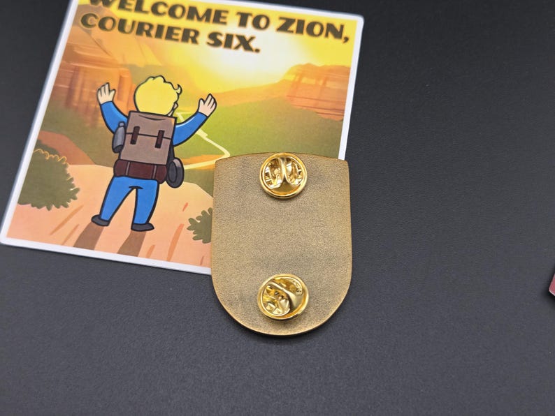 May include: A gold-colored metal pin with two pin backs. The pin is shield-shaped. A card with the text "WELCOME TO ZION, COURIER SIX" and a cartoon character is next to the pin.
