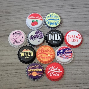 May include: A collection of ten circular metal bottle caps featuring various Nuka-Cola themed designs. The caps display colorful graphics and text including Nuka-Cola, Nuka Twist, Nuka Grape, Nuka Dark, Nuka Cherry, Nuka Wild, Nuka Orange, Nuka Victory, and Nuka Quantum.