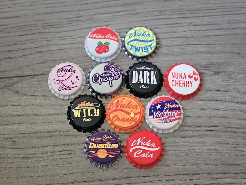 May include: A collection of ten circular metal bottle caps featuring various Nuka-Cola themed designs. The caps display colorful graphics and text including Nuka-Cola, Nuka Twist, Nuka Grape, Nuka Dark, Nuka Cherry, Nuka Wild, Nuka Orange, Nuka Victory, and Nuka Quantum.