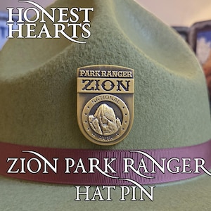 Może przedstawiać: Zielony kapelusz z bordową wstążką i złotą przypinką. Na przypince widnieje napis "Park Ranger Zion National Park" i znajduje się na niej wizerunek góry. Na kapeluszu widnieje napis "Honest Hearts", "Zion Park Ranger" i "Hat Pin".