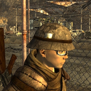 May include: A person wearing a tan safari hat with a dark brown band and a small white patch. The person is also wearing glasses and a brown scarf. A rifle is visible in the background.