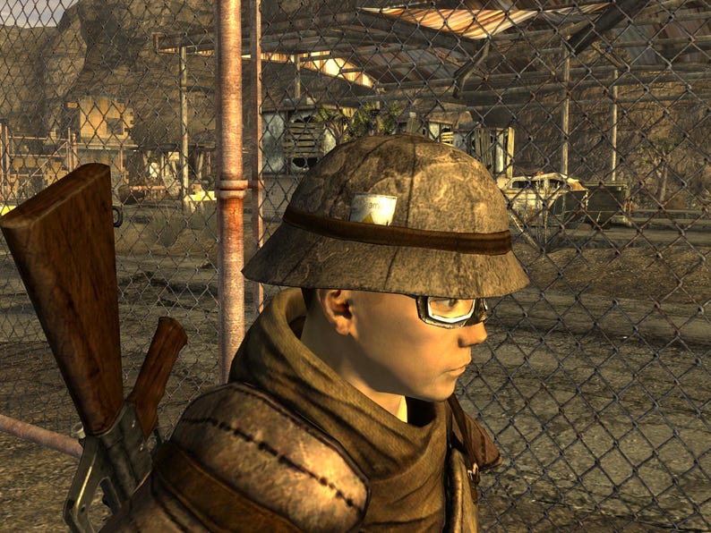 May include: A person wearing a tan safari hat with a dark brown band and a small white patch. The person is also wearing glasses and a brown scarf. A rifle is visible in the background.