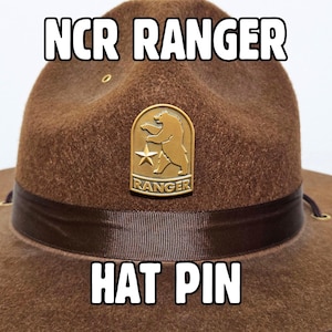 May include: A brown hat with the text "NCR RANGER HAT PIN" in white block letters. The hat features a gold-coloured pin with a bear and star emblem. The hat has a dark brown band.